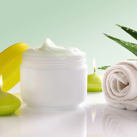 White plastic container with facial or body cream of aloe vera. Candles, towel and plant decoration and green background isolated. Front view